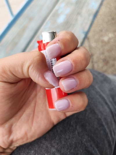 A close-up, overhead shot shows a person's hand holding a red lighter with a silver top. The person's fingernails are painted a light, milky pink. The background consists of a wooden bench with blue accents and a gravelly surface. The scene is located in the city of Borodino, Russia.