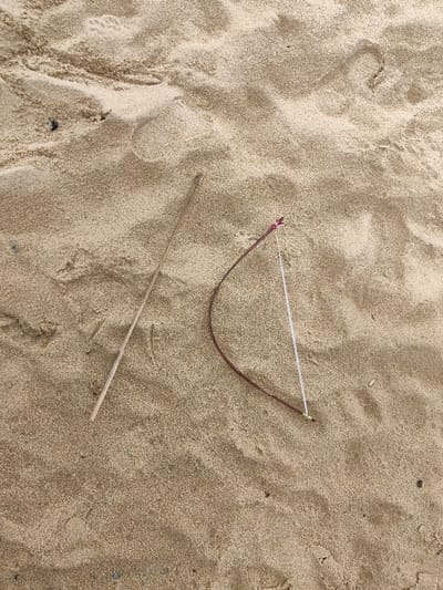 The scene, located in Thuận An Ward, Vietnam, displays a granular, light brown sand surface marked by subtle undulations and shallow impressions. Two distinct objects rest on the sand. On the left, a straight, slender stick, light tan in color, lies diagonally across the frame. To its right, a curved, dark reddish-brown object, consistent with a flexible twig or root, has a white string tautly stretched between its two ends. The string is secured at the object's upper terminus by a small, dark reddish binding, and at its lower terminus by a small, yellowish or light-colored knot. Scattered dark particulates are visible across the sand. No individuals are present, and no actions or interactions are depicted.
