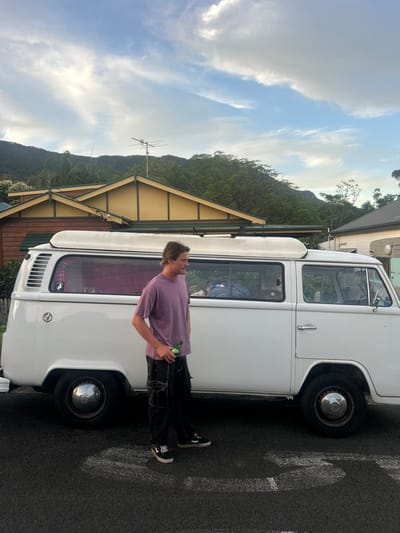 A young male stands beside a white Volkswagen Type 2 (Bay Window) camper van on asphalt pavement in Thirroul, Australia. The male has medium-length light brown hair, wears a faded purple t-shirt, dark cargo pants with visible white stitching, and black and white sneakers. He holds a green glass bottle in his right hand and is looking towards his right, out of frame. The van features a collapsed pop-top roof, a roof rack above the driver's side, and chrome hubcaps; a red curtain and assorted items are visible through its windows. In the background, two single-story residential buildings are present: one with a brown timber facade and a yellow gable roof featuring dark green trim, surmounted by a television antenna; another building, possibly a house or trailer, is partially visible on the far right. Behind the structures, the dense, tree-covered slopes of the Illawarra Escarpment rise under a partially cloudy blue sky. Faint white markings are visible on the asphalt in the foreground.
