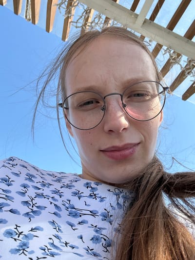 The image shows a close-up, low-angle selfie of a young woman with blonde hair and freckles, wearing glasses and a white dress with a blue floral pattern. She is looking towards the camera with a slight smile. The background consists of a clear blue sky and a wooden structure overhead, possibly part of a canopy or pergola. The lighting suggests it is daytime, likely outdoors. The location is identified as Металлплощадка, Russia.