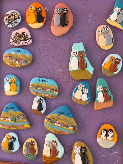 The image displays a collection of decorative painted stones arranged on a purple surface. The stones are designed to resemble animals, primarily cats and owls, and some depict landscapes with mountains and villages. Several of the landscape stones are inscribed with the word "Чифлик" (Chiflik), indicating they are related to the town of Chiflik, Bulgaria. The craftsmanship involves small, rounded stones painted with bright colors and distinct details. There are no people or discernible actions occurring in the scene. The overall arrangement suggests a display of handmade crafts or souvenirs.
