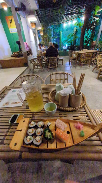 The scene depicts a restaurant setting in Phan Rang – Tháp Chàm, Vietnam. A bamboo table is laden with a wooden boat-shaped tray filled with assorted sushi, including maki rolls and nigiri, accompanied by pickled ginger and wasabi. There's a pitcher of liquid, possibly tea or juice, and a set of tea cups with chopsticks in a bamboo holder. A menu is also visible on the table. In the background, people are seated at other bamboo tables and chairs, with a view of the outdoor setting including a fence, foliage, and overhead lighting. A standing fan is visible in the lower left corner.