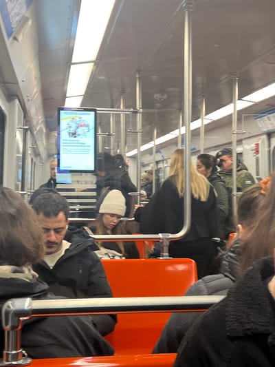 The image depicts the interior of a Helsinki Metro M100 series train car, operating within the city of Espoo, Finland. The environment features white walls and ceiling with integrated fluorescent lighting, orange plastic seating, and numerous silver vertical support poles and horizontal grab bars. A digital display screen mounted from the ceiling shows a metro line map and Finnish text, prominently featuring "HELSINGIN METRO" at the top. A security camera is visible on the ceiling in the mid-ground.

Approximately 15-20 individuals are present. In the mid-ground, a man with a mustache and dark hair is seated on an orange seat, wearing a black puffer jacket over a light shirt, with a white earbud in his right ear, looking downwards. To his right and slightly behind him, a young woman with long dark hair wears a cream-colored beanie and a dark jacket, also looking downwards. Standing in the central aisle, a woman with long blonde hair, dressed in dark clothing, faces right. Behind her, a man in a camouflage jacket and a dark blue beret with an emblem is visible, facing right. Other passengers, mostly in dark winter attire, are visible seated and standing throughout the car, many partially obscured. No direct interactions between individuals are clearly observable; most appear to be independently occupied.