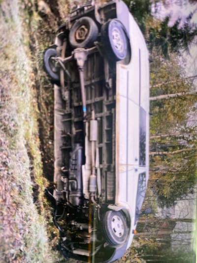 The image shows an overturned white van in a wooded area. The van is resting on its side with its wheels facing upwards. The undercarriage of the van is visible, including the exhaust system and suspension components. The surrounding environment consists of trees and undergrowth, suggesting a rural or semi-rural location. The image is taken from a slightly elevated perspective, looking down at the van. There is no indication of the specific location within the city of Kerava, Finland. No people are visible in the image, and no actions or interactions are depicted. The scene appears to be a static representation of a vehicle accident.