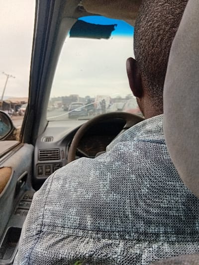 The scene is observed from inside a vehicle, looking forward through the windshield in Itobe, Nigeria. A dark-skinned individual, visible from the back of their head and right ear, occupies the driver's seat, wearing a light-colored short-sleeved shirt patterned with a black geometric design. The driver's hands are positioned near a dark steering wheel.
The vehicle's interior components include a visible dashboard with air vents and controls, and a left door panel equipped with power window switches and a worn armrest. The windshield displays a distinct horizontal crack across its upper portion and a blue tint strip at the top edge.
Externally, an overcast sky is visible above a paved road populated with multiple vehicles, indicating active traffic. On the left side of the road, various low-rise structures, consistent with stalls or small businesses, are present, alongside utility poles.