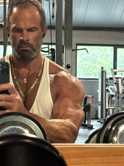 The image depicts a muscular man in a gym, taking a selfie in a mirror. He is wearing a white tank top and multiple gold necklaces. His left arm, which is prominently displayed, shows well-defined muscles and veins. In the background, gym equipment, including weight racks and exercise machines, is visible. Large windows offer a view of greenery outside, suggesting the gym is in an area with natural surroundings. The man has a beard and short hair, and he is wearing black earbuds. The foreground of the image is dominated by the reflection of gym weights in the mirror. The overall impression is one of a dedicated fitness enthusiast in a typical gym setting. The location specified is Jemelle, Belgium.