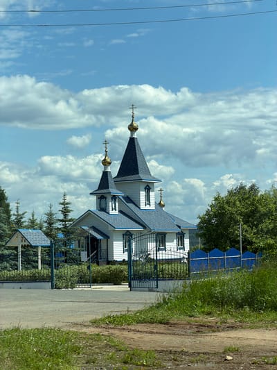 This is a daytime, eye-level view of a white and blue Eastern Orthodox church situated in Кварса, Russia, under a bright blue sky filled with numerous white cumulus clouds.

The church, constructed with white horizontal siding, features a dark blue shingled roof and prominent blue trim around its windows and architectural elements. It has two main spires, each topped with a conical dark blue roof, a small golden onion dome, and a golden Orthodox cross. A third, smaller golden onion dome with a cross is visible on a lower roof section towards the left. The church entrance, located centrally, includes a small porch with a blue roof and dark metal railings.

The church grounds are enclosed by an ornate dark blue or green metal fence. A matching, decorative metal gate stands open in the foreground, providing access to the property. To the left of the gate, within the fenced area, is a small, white A-frame structure with a blue shingled roof, resembling a shrine or canopy. Several young, conical coniferous trees are planted around the church, and mature green deciduous trees are visible on the right side, behind a corrugated blue metal fence.

The immediate foreground consists of a dirt or gravel road, showing patches of green grass and small puddles or damp areas. Two dark, thin power lines stretch horizontally across the top of the sky. No individuals are present in the scene.