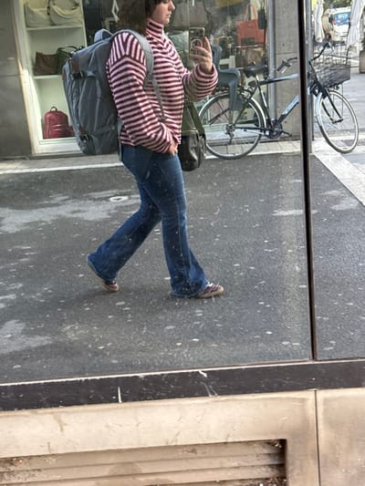 An individual is reflected in a storefront window on an urban street in Arezzo, Italy. The person, viewed from the left side, walks from left to right across the frame. They are wearing a pink and maroon horizontally striped turtleneck sweater, blue flared jeans splattered with white specks, and dark red sneakers with tan soles. A large grey backpack with brown accents is worn on their back, and a dark green shoulder bag hangs on their left side. Their right hand holds a light-colored smartphone, raised in front of them, possibly capturing a selfie or video. A ring is visible on their right ring finger.
The reflective surface shows the street in the foreground, characterized by dark grey pavement with numerous white speckles, likely water or residue. A black horizontal bar at the bottom marks the window's base, below which is a light-colored, possibly concrete, structure with horizontal slats.
Behind the reflection, the interior of a store is visible through the window on the left, displaying various bags, including a light grey tote, a dark brown shoulder bag, and a red backpack on white shelving. To the right, on what appears to be a sidewalk, two dark-colored bicycles are parked: one with a black child seat on the rear rack and a silver front fender, and another further right with a front basket. A white vehicle is partially visible in the far background beyond the bicycles.