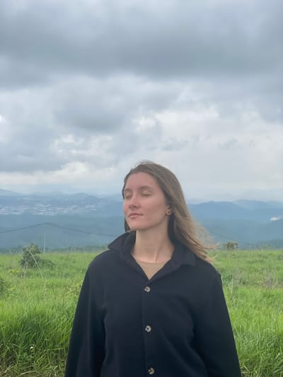 A young woman with her eyes closed stands in a grassy field, facing the camera. She is wearing a black collared jacket over a tan shirt. Her hair is long and brown, and it is being blown by the wind. In the background, there are rolling hills and a city in the distance. The sky is cloudy and gray. The scene is likely located in the city of Trường Thọ, Vietnam, given the geographical context of rolling hills and a visible city in the distance.