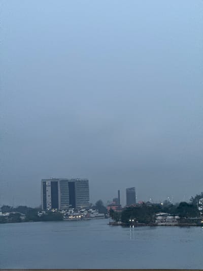 The image displays an urban waterfront scene in Falomo, Lagos, Nigeria, characterized by an overcast, light blue-grey sky occupying the upper two-thirds of the frame. The lower third features a calm body of water, likely the Falomo Lagoon.

Across the water, on the distant shoreline, significant urban development is visible. Prominently positioned on the left is a modern, multi-story building complex, identifiable as the Oando Wings Office Complex. It consists of two tall, dark-glassed towers with vertical panels, connected by a lower structure. A yellow logo is discernible near the top of the left tower. Below this complex, closer to the water's edge, are several smaller, illuminated structures.

Towards the center-right of the distant shore, additional multi-story buildings are present. These include a tall, slender, dark vertical structure, possibly an industrial stack or chimney, and another building with a distinctive angled or sloped roofline to its right. Interspersed among the structures are patches of green vegetation, primarily trees and shrubs, particularly on the landmasses closest to the foreground and along the distant banks. Numerous small, artificial lights are illuminated on the buildings and along the waterfront, indicating low ambient light conditions, suggesting either dusk, dawn, or heavy cloud cover. No individuals, vehicles, or specific human activities are discernible within the frame.