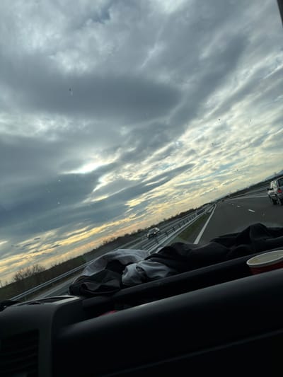 The photograph shows a view from inside a vehicle, likely a car, traveling on a multi-lane highway. The highway has a median barrier. Other vehicles are visible on the road. The sky, filled with numerous clouds, dominates the left portion of the frame and appears to be reflected in the vehicle's windshield. The interior of the car is partially visible on the right, with clothing and a cup placed near the dashboard. The image was not taken in Vodenichane, Bulgaria; the highway environment indicates a more developed infrastructure not typically found in smaller villages.