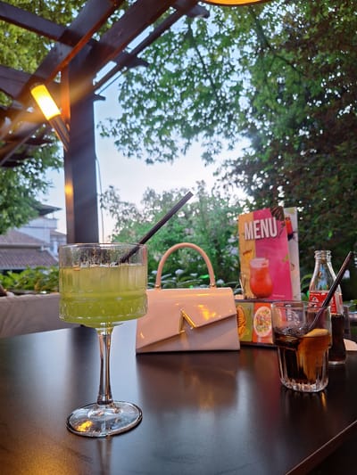 This image depicts an outdoor dining or lounge area in the city of Petrich, Bulgaria. The primary focus is on a dark, reflective table displaying two beverages, a handbag, and a menu stand.

On the left, a tall, stemmed glass contains a clear, light-green liquid with a black straw. The glass features a ribbed pattern on its lower bowl. To its right, a light-colored, structured handbag with a gold-toned geometric (appearing as an 'A' or 'V') clasp rests on the table. Further to the right, a shorter, wider glass contains a dark brown liquid, ice, and a lemon wedge, also with a black straw. Adjacent to this glass is a small, clear glass bottle of Coca-Cola with a red label. A colorful menu or promotional stand, featuring the word "MENU" prominently, an image of an orange drink, and other food items, stands behind the handbag and between the drinks.

The background reveals an outdoor setting. Dark-colored structural beams, likely part of a pergola or awning, span the upper portion of the image. A glowing, cylindrical yellow-orange lamp is affixed to one of these beams on the upper left. Dense green foliage, characteristic of trees and bushes, fills the midground and background, with patches of bright sky visible through the leaves. On the far left, beyond the foliage, glimpses of residential or commercial building structures with rooftops can be seen. No individuals are visible in the scene. The overall lighting suggests either late afternoon or early evening.