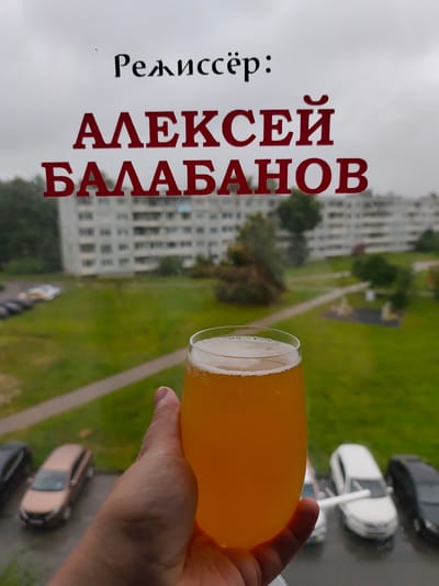 A close-up, slightly angled shot captures a hand holding a glass of light orange, frothy beer. The glass is positioned in the foreground, obscuring a portion of the background. Through the glass, a city scene is visible, appearing slightly out of focus. The background depicts a grey, overcast sky, a multi-story apartment building with many windows, a green grassy area with trees, a paved pathway, and several parked cars including a light-colored sedan, a white sedan, and a black sedan. The text "Режиссёр: АЛЕКСЕЙ БАЛАБАНОВ" is superimposed on the image, with "Режиссёр:" in black serif font and "АЛЕКСЕЙ БАЛАБАНОВ" in red block letters. The presence of the apartment building, cars, and the general urban landscape suggests the location is within a city or town. Based on the text, this image is likely related to director Alexei Balabanov and may have been taken in or represent a scene connected to his work or life in the city of Syaskelevo, Russia.