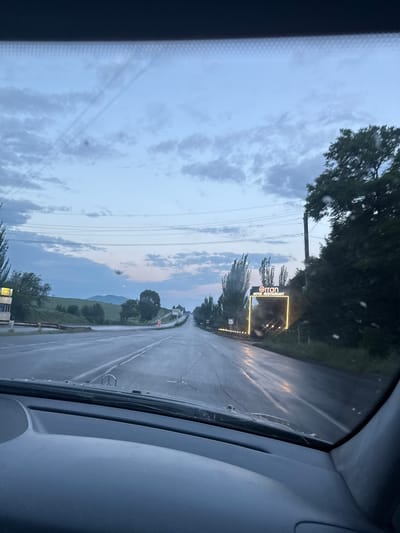 The image, taken from inside a vehicle looking out through the windshield, depicts a road scene at dusk. The road is wet, suggesting recent rain, with reflections of lights visible on its surface. Overhead power lines stretch across the sky, which is filled with a mix of clouds and a pale blue twilight.

On the right side of the road, a distinctive illuminated sign is visible. The sign is rectangular with rounded corners, outlined in bright yellow light, and displays the text "Orran HOTEL RESTAURANT" in dark letters against a black background. The illuminated outline of the sign and a string of smaller lights running along the base of the sign create a warm glow. Trees, with dark foliage, flank the road on the right.

Further down the road, on the left, there is a small building with yellow signage, possibly a kiosk or a shop. Rolling hills covered in greenery form the background, with a faint outline of mountains visible in the distance. The overall impression is of a rural or semi-rural highway at the end of the day. The vehicle's dashboard is partially visible in the foreground.