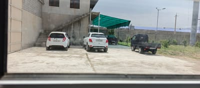 The image shows a scene in Mehrabad, Uzbekistan. In the foreground, on a paved surface, are two white passenger cars. The car on the left is a Chevrolet Spark, with license plate 30 T 804 JB. The car on the right is a white SUV, possibly a Chevrolet Trax, with license plate 30 J 013 SB. To the right of the white SUV is a dark-colored sedan, partially obscured by the SUV, with license plate 30 J 376 CHR. Further to the right and slightly behind the white SUV is a black pickup truck with a flatbed, license plate 30 L 795 QB.
Behind the vehicles, a concrete building with external stairs is visible. The stairs lead up to a structure with a green canopy. To the right of the building, a long brick wall encloses an area with sparse trees and grass. Beyond the brick wall, there are buildings with blue roofs and a tall concrete structure. The sky is overcast with some visible clouds. The overall impression is of an outdoor parking area or industrial/commercial space.