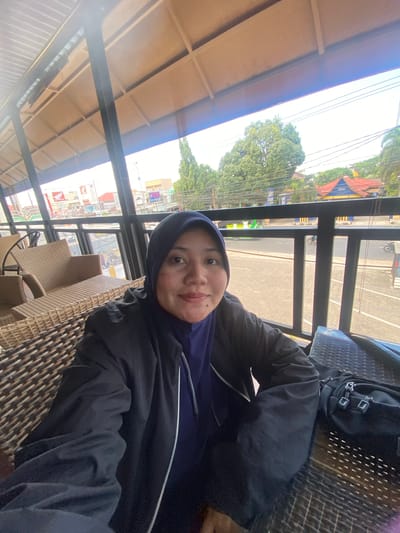 A woman wearing a hijab and a dark jacket is seated at a table, likely outdoors or on a balcony, with a view of a street in Keraton, Indonesia. She is looking towards the camera with a neutral expression. In the background, there are buildings, trees, and a street with some traffic, including motorcycles. The seating area appears to be made of wicker or rattan furniture, and there is a black bag on the table next to her. The lighting suggests it is daytime.