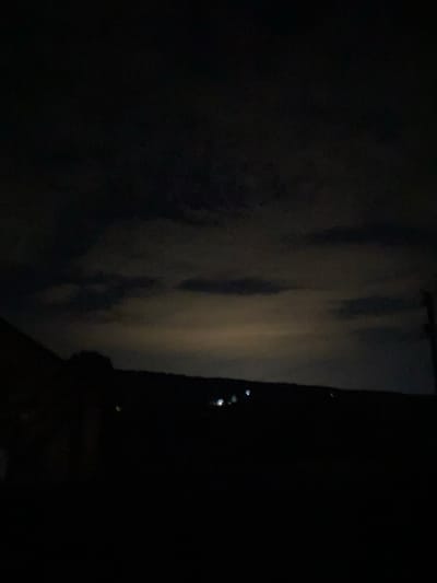 This is a dark, horizontally oriented photograph taken at night in Negrești, Moldova. The scene is dominated by deep shadows, with only faint hints of detail visible. On the left side of the image, a dimly lit, vertical surface appears to be a wall or curtain, illuminated by an unseen light source that creates soft, diffused highlights and shadows. This surface appears textured, with vertical lines and mottled patches suggesting fabric or a textured wall.
To the right of this illuminated surface, the image transitions into complete darkness. However, within this blackness, a scattering of small, bright points of light are visible. These lights are irregular in shape and intensity, and their arrangement suggests they could be distant city lights, stars, or perhaps lights from a vehicle or building. The overall impression is of looking out into a dark night from a slightly illuminated interior or sheltered space.
The lack of clear subjects or discernible objects makes it difficult to pinpoint a specific activity or event. The time of day is clearly night, and there are no strong indicators of weather conditions, though the darkness and the diffused light could suggest a cloudy or overcast night. The presence of scattered lights in the distance hints at a populated area, possibly a town or city outskirts. The overall mood is mysterious and somewhat subdued due to the low light conditions. There is no visible text in the image.