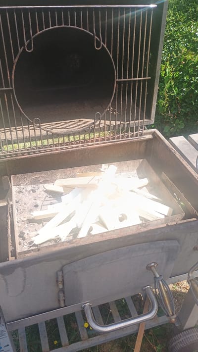 The image shows an outdoor grill, likely in Windeck, Germany, with its lid open. The grill has a large, circular chamber at the top, which appears to be a smoker, and a lower rectangular compartment. The lower compartment contains a pile of what appears to be light-colored wood or kindling, resting on a metal grate. There are remnants of ash and debris in the bottom of this compartment, suggesting recent use or preparation for a fire. The grill itself is made of metal, showing signs of rust and wear. The background consists of green foliage, indicating an outdoor setting with vegetation. The lighting suggests it is daytime, with sunlight creating some glare on the grill's surface. No people are visible in the image, and there are no ongoing activities.