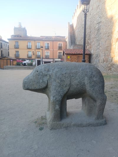 The image shows a stone sculpture of a stylized animal, likely a bull, in the foreground. The sculpture is positioned on a sandy ground surface in what appears to be a public square. In the background, there are several multi-story buildings with tiled roofs and balconies, and a section of a thick stone wall with battlements, characteristic of ÁVila, Spain. A lamppost stands to the right of the sculpture, and a small wooden structure with a tiled roof is also visible near the stone wall. The sky is clear and bright, suggesting it is daytime.