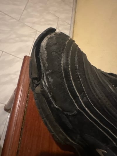 An extreme close-up, eye-level indoor scene in Macedo de Cavaleiros, Portugal, depicts a single, severely degraded black athletic shoe. The shoe rests on a dark brown, polished wooden surface, which exhibits a visible grain and a vertical seam along its left edge. To the left of the wooden surface, a light-colored, possibly off-white or light gray rectangular tiled floor is visible, arranged in a staggered pattern with dark grout lines. The background on the upper right features a plain, light yellow wall.
The shoe's primary subject is its rear-right quarter. The black sole is extensively delaminated, cracked, and degraded, particularly at the heel and along the side. Deteriorated material appears chalky white in areas where the sole has separated or worn away. The upper part of the shoe is black with several prominent, parallel, light-colored stripes running diagonally across it, consistent with a synthetic material. A faint, lighter-colored emblem is partially visible on the lower right section of the shoe's upper. No individuals are present in the image, and no actions or interactions are depicted. The scene is static, illuminated by ambient indoor lighting.