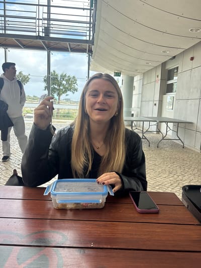 An interior scene in a modern building located in Porto Salvo, Portugal, depicts a young adult female seated at a brown wooden table, smiling directly at the camera with her mouth slightly open. She has long blonde hair and is wearing a black v-neck top and a black leather-like jacket, accessorized with gold hoop earrings and a gold pendant necklace. Her fingernails are painted red. She holds a silver fork upright in her right hand, while her left hand rests near a clear plastic food container with a blue lid, which contains a meal. To her right on the table is a purple-cased smartphone. In the background, a young adult male in a white hoodie and light-colored pants walks left, facing away from the camera, with a dark bag slung over his shoulder. The room features a light brown cobblestone floor, light grey paneled walls with visible fasteners, and a white ceiling that includes a curved, paneled section and exposed metal beams and glass windows. Through the large windows, an outdoor environment with wind-swept trees and an open landscape under an overcast sky is visible. Two grey folding tables are stacked against a far wall on the right side of the room.