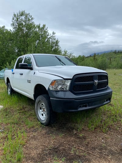 This is a media file of a white Dodge Ram 1500 pickup truck parked on a grassy area with some dirt and dry vegetation. The truck is facing slightly towards the right side of the frame, with its front end visible. It has a black grille with a chrome Ram logo and black bumper. The headlights are clear, and the windshield has wipers. The side of the truck shows a "Ram 1500" badge. One of the front wheels and one of the rear wheels are visible. The truck appears to be parked in an outdoor setting, with a background of lush green trees and a cloudy sky. In the distance, there are mountains visible through the trees. The overall impression is a slightly overcast day, possibly in a rural or wilderness area. There are no people visible in the image. The scene appears to be in Haines Junction, Canada, indicated by the location context. The weather appears to be cloudy and possibly damp, as there are water droplets visible on the truck's surface. The ground is covered in grass and dirt. No specific activity is taking place; the truck is simply parked.