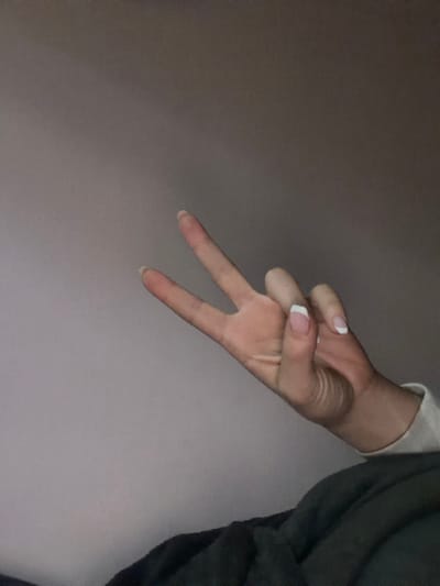 The image displays a close-up of a human left hand, angled from the lower right toward the upper left. The hand has light skin tone and exhibits a French manicure on the visible fingernails (thumb, index, middle, and ring finger). The index and middle fingers are extended upwards in a "V" shape, while the thumb, ring, and pinky fingers are curled towards the palm. A portion of a light-colored sleeve or cuff is visible on the wrist area of the hand. In the lower right foreground, a dark, textured fabric object, possibly a blanket or clothing, is present. The background is a plain, solid wall, colored a muted grayish-purple, suggesting an indoor environment. No elements within the image provide geographical context to determine the location as Obruchishte, Bulgaria, or any other specific city or country.