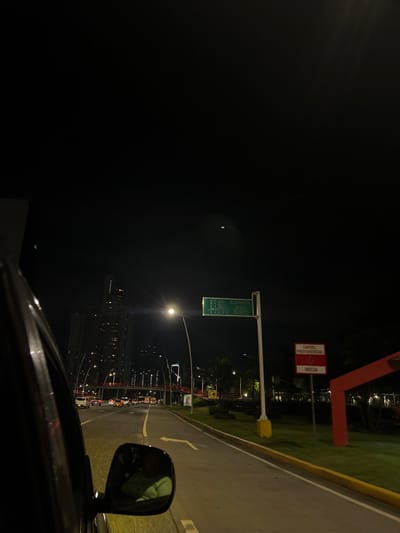 This nighttime image depicts an urban roadway scene in Panama City, Panama, observed from the passenger side of a moving vehicle. The sky is dark, illuminated by artificial light sources.
In the immediate foreground on the left, a portion of the vehicle's dark door frame and side mirror are visible. The side mirror contains a distorted reflection of an individual wearing a light-colored top.
The roadway ahead is a multi-lane thoroughfare with moderate vehicle traffic, indicated by numerous illuminated headlights and taillights. Road markings include solid yellow lines separating traffic and dashed white lane lines. A white arrow is painted on the pavement, indicating a turn.
To the right of the road, a tall pole supports a green overhead street sign with white lettering. The sign displays directional information, including "PTO. CARN.", "SANTA ANA", "CALIDONIA", "507", "BALBOA", and "DOBO". Below this, a red and white sign reads "CARRIL PREFERENCIAL INICIA" (Preferential Lane Starts). A bright streetlight on a curved pole illuminates this area. Further down the road, a distinctive red pedestrian overpass structure spans the width of the road.
The background features a dense collection of tall, illuminated skyscrapers, characteristic of Panama City's modern skyline. The scene is located on a major urban artery, consistent with Avenida Balboa or Cinta Costera, specifically in an area leading towards central districts like Santa Ana and Calidonia.