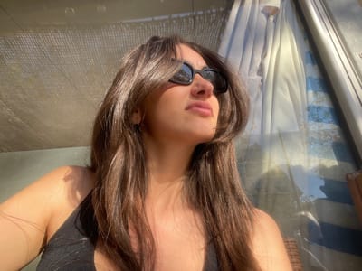 A single female subject is depicted in a sunlit, close-up, angled portrait, situated in what appears to be a shaded outdoor or semi-outdoor area in La Grande-Motte, France. She has long, dark brown hair, wears black sunglasses, and a black sleeveless top. Her head is tilted upwards and her gaze is directed towards the upper right, with her lips slightly parted. Strong natural light illuminates her right side. The background on the left features a woven, translucent light-colored fabric, likely a canopy or sunshade. The right background shows a reflective glass surface, such as a window or door, displaying reflections of light-colored curtains with blue stripes and indistinct outdoor elements. A segment of light-brown woven furniture is visible on the far right.