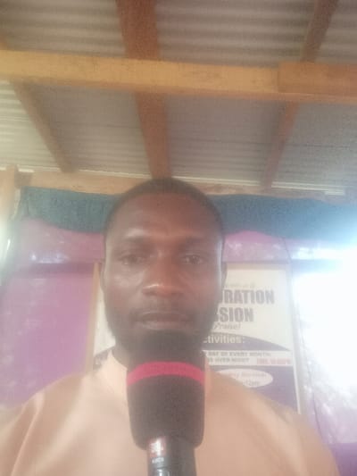 The image depicts a male individual positioned centrally, from the chest up, within an indoor setting in Nara Unateze, Nigeria. The man, with dark skin, short dark hair, and a beard, wears a light peach-colored collared shirt and looks directly forward. A black microphone with a red band near its top, featuring "JBL" and "KMC6" branding, is held or positioned in front of him, obscuring the lower portion of his face and chest.
Behind the man, a white sign or poster is visible, partially obscured and blurred. Identifiable text fragments include "...up with us @", "RATION SSION", "(Praise)", "Activities:", "DAY OF EVERY MONTH:", "OVER-NIGHT TIME: 10:00PM", and "Sunday Services:". The background immediately behind the sign consists of a purple wall, with a dark blue or green fabric draped horizontally across the upper part. Above the individual and background, the ceiling structure is visible, comprising corrugated metal sheets supported by wooden beams. Light appears to be originating from the right side of the frame.