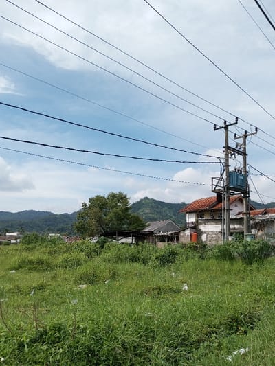 The scene depicts a semi-urban or rural landscape in Kandenuwara, Sri Lanka, under a partly cloudy sky. The foreground consists of an overgrown field of tall green grass, interspersed with scattered white plastic debris. In the midground, a prominent concrete utility pole stands, supporting multiple cross-arms, insulators, and numerous black electrical power lines traversing the frame horizontally. A blue electrical transformer is mounted on this pole. To the right of the pole, a multi-story building with white and exposed concrete walls, a reddish-brown tiled roof, and visible windows is present. An orange cylindrical tank is attached to its exterior. To the left, a smaller single-story structure with a dark roof is partially visible, flanked by mature trees. In the background, rolling green, forested hills rise, some displaying terraced sections. The scene is devoid of human or animal subjects, depicting a static environment under bright daylight conditions.