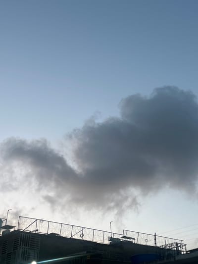 The image captures a cloud formation against a pale blue sky, partially obscuring a rooftop structure. The rooftop is equipped with a dark metal fence and various objects, including what appears to be lighting fixtures. The overall lighting suggests either early morning or late afternoon. The scene is situated in the urban environment of Conuco Viejo, Venezuela.