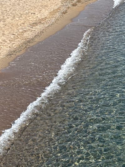 This is an aerial view of a sandy beach meeting clear, shallow water. The sand is light brown and textured with small pebbles and debris. A gentle wave is breaking on the shore, creating a line of white foam. The water is transparent, revealing the sandy bottom and the patterns created by the sunlight reflecting off the ripples. The scene is serene and natural, evoking a sense of tranquility. There is no visible text. The time of day appears to be sunny, as indicated by the bright reflections on the water. The location is Koshkol', Kyrgyzstan.