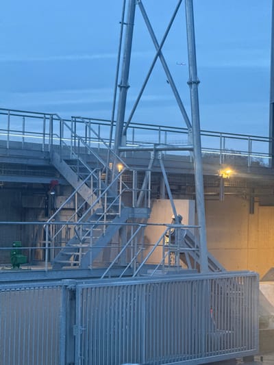 A tall metal structure with industrial stairs rises against a light blue sky. The framework includes crisscrossing supports, a grated platform, and a series of steps leading to different levels. A small bird flies near the top. A building or structure is visible in the background with faint lights on. Parts of machinery and metal gratings are seen in the bottom.