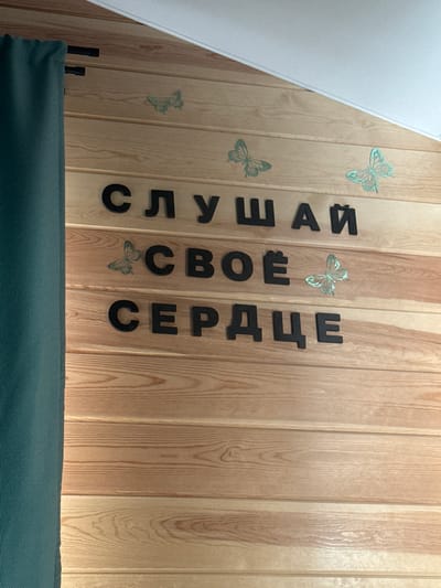 The image shows a wooden wall with black lettering spelling out "СЛУШАЙ СВОЁ СЕРДЦЕ" which translates to "LISTEN TO YOUR HEART" in Russian. There are several decorative butterflies placed on the wall above and between the words. The wall is made of horizontal wooden planks, and a teal curtain is visible on the left side of the image, obscuring part of the wall and the full extent of the lettering. The scene appears to be indoors, possibly in a room or establishment in Khuzhir, Russia.