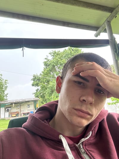 A medium shot depicts a person with short, dark hair wearing a maroon hoodie. The person's right hand is touching their forehead. The background shows a green lawn, trees, and a building that appears to be a trailer or modular structure. A partial view of scaffolding or a similar structure is in the upper left corner. The location is Dabovets, Bulgaria.