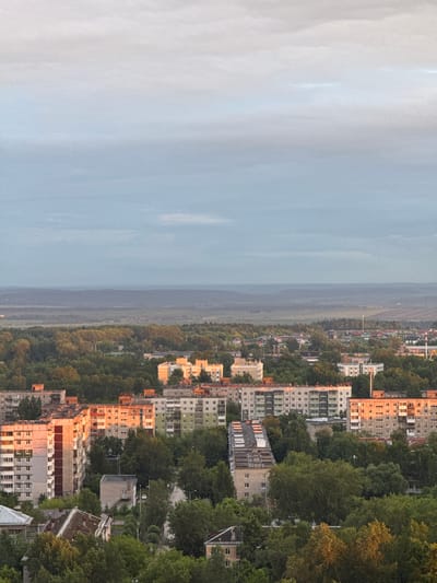 This image presents a detailed forensic view of a cityscape within Kitsjanovo, Russia, observed under an overcast sky, likely during sunrise or sunset due to specific lighting conditions. The upper third of the frame shows a gradient sky, transitioning from a pale blue-grey at the bottom to a lighter, almost white-grey at the top, with indistinct cloud layers.

Below the sky, the distant horizon reveals undulating landforms, appearing as dark blue-grey masses, consistent with forests or low hills, obscured by atmospheric haze. The mid-ground is dominated by a vast expanse of green vegetation, primarily dense tree cover, interspersed with scattered residential buildings and what appear to be agricultural fields in the far distance.

The foreground and mid-ground are populated by a dense urban residential area. Numerous multi-story apartment buildings, characteristic of post-Soviet architectural styles (e.g., concrete panel construction), are visible. Their facades range in color from light grey and off-white to structures with darker vertical accents. Crucially, several buildings, especially on the left and right sides of the lower frame, are strongly illuminated by a warm, orange light, indicating a low-angle sun. This light contrasts with the generally muted tones of shadowed buildings and the sky.

Extensive mature green foliage, consisting of numerous trees, is integrated throughout the urban landscape, obscuring ground-level details, roads, and smaller structures. Glimpses of dark grey paved roads are visible winding through the greenery and between buildings. No individuals, specific actions, or interactions are discernible within the frame. The scene is static, capturing a wide panoramic view of a residential area heavily integrated with natural elements under diffused natural light, with specific areas highlighted by direct, low-angle sunlight.