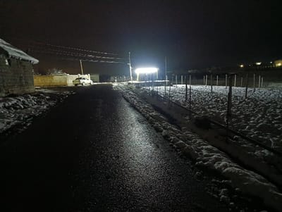 A snow-dusted landscape at night. A dark, wet road leads into the distance, illuminated by a bright light source ahead. A white car is parked near a wall on the left. To the right of the road, snow covers the ground, and a structure with vertical posts is visible. Houses or buildings are vaguely visible in the far distance, and overhead wires stretch across the sky. The night is dark, and the snow adds a bright contrast to the scene.