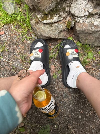 The image shows a person's legs and feet, wearing black slides with white socks, holding a bottle of Corona Extra beer with a lime wedge. The scene is outdoors, with gravel, grass, and large rocks. The environment appears to be natural, possibly a park or trail. There is no indication of Cheposh, Russia.