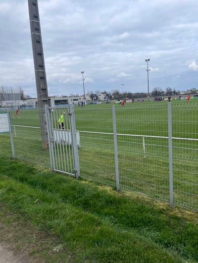 This image depicts an outdoor football (soccer) pitch in Oberschaeffolsheim, France, under an overcast sky. In the foreground, a strip of natural grass and soil leads to a grey galvanized wire mesh fence, approximately 1.5 meters in height, featuring an open single-leaf gate with vertical metal bars. A tall, segmented concrete utility pole stands prominently on the left side, partially obscuring the view. Beyond the fence, on the green turf playing field marked with white lines, multiple individuals are observed. Several players are dressed in red kits, while others wear dark blue kits, indicating two distinct teams engaged in a game or practice. One individual, wearing a high-visibility yellow bib over dark attire, is visible through the open gate, standing on the field. In the background, beyond the pitch, a mix of buildings, including residential structures and potential industrial facilities, is visible. A line of trees borders the horizon, and two prominent floodlight towers with multiple lamps illuminate the field from the rear. Advertising banners in purple and red are visible along the far sideline of the pitch.