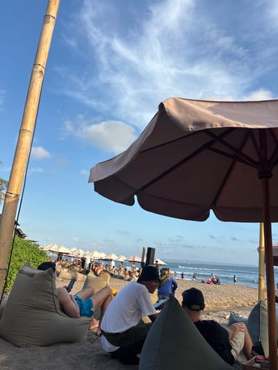 This image captures a serene beach scene in North Kuta, Indonesia. The foreground is dominated by large, comfortable beanbag chairs set on the sandy beach. Several people are seated on these chairs, some engaged with their phones, others simply relaxing and enjoying the view. Behind them, a line of beach umbrellas offers shade to more beachgoers scattered along the coastline. The ocean stretches out to the horizon, with gentle waves and a few distant figures in the water. The sky above is a clear blue, dotted with wispy clouds, indicating pleasant weather. A tall bamboo pole stands prominently in the left foreground, adding a natural element to the composition, while a large umbrella partially obscures the view in the right foreground. The overall atmosphere is one of leisure and tranquility, typical of a popular beach destination.