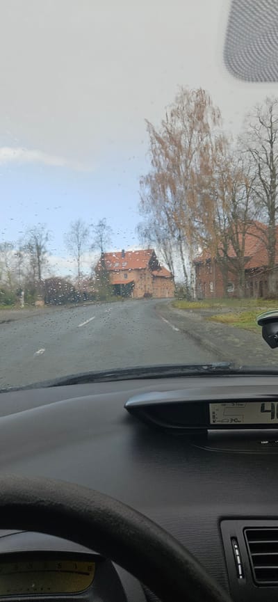 The image shows a view from inside a vehicle, likely a car, on a rainy day. The landscape outside the windshield is blurred due to the rain droplets. There are buildings with reddish-brown roofs visible, and trees with bare branches, characteristic of late autumn or early spring. The buildings are typical of rural or suburban architecture. Based on visual similarity and the request, these buildings are likely in Delbrück, Germany. The dashboard of the car is visible, including the steering wheel, a digital display showing vehicle data, and part of the air conditioning vents. The angle is rotated, with the outside world appearing sideways.