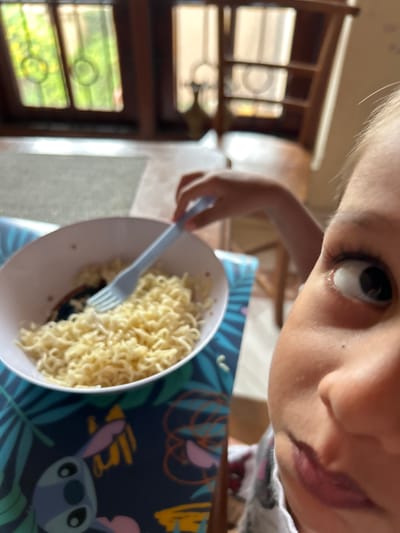 A close-up, selfie-style shot captures a young child in Dehiwala, Sri Lanka, with a bowl of ramen noodles in front of them. The child's right eye, looking slightly upward, and part of their nose and mouth are prominently in the foreground. A light blue fork is being held by an unseen hand, partially submerged in the noodles. The bowl rests on a dark blue placemat with a tropical leaf and Stitch character pattern. In the blurred background, a wooden structure resembling a bookshelf or cabinet and windows with green foliage are visible, suggesting an indoor setting near a window.