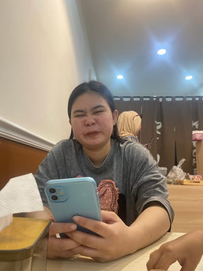A young East Asian woman with dark hair is captured in a medium shot, seated at a table in what appears to be a restaurant. She is holding a light blue smartphone in her hands, her fingers delicately positioned on the screen. Her expression is one of humorous contortion, with her eyes squeezed shut and her mouth puckered in a playful grimace. She wears a grey t-shirt with a pink graphic design on the front. To her left, a white paper napkin dispenser is visible on the wooden table. In the background, other patrons are present, including a woman wearing a beige hijab, also looking at her phone. The environment suggests an indoor setting, possibly with the warm glow of overhead lights, which reflect off the ceiling. The walls are a neutral tone, and brown curtains hang in the background, suggesting a partitioning of space within the establishment. The scene does not provide any specific visual cues to pinpoint the exact location within Watampone, Indonesia, beyond it being an indoor dining area.