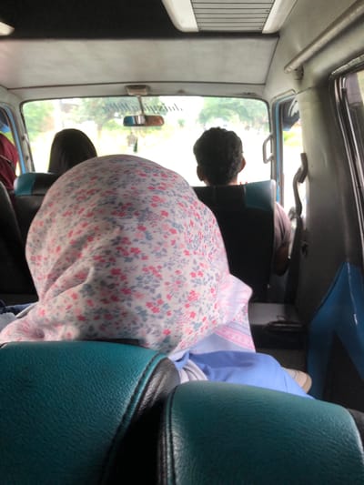 The image, taken in what appears to be a moving vehicle, perhaps a small bus or van, shows the interior of the vehicle with passengers. The scene is dominated by the back of a person's head, covered by a hijab-like garment with a small floral pattern, possibly a pink and blue design. To the right of this person is a blue seat, likely vinyl or leather. The vehicle's windows offer a blurred view of the outside, potentially showing trees or vegetation. There is also another person further up in the bus but its hard to see details. The image was captured in Putoh Sa, Indonesia.