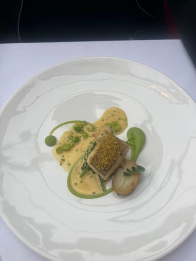 A high-angle, close-up photograph captures a single, artfully plated dish on a large, round, white ceramic plate within a dining establishment in Liverpool, United Kingdom. The plate exhibits subtle concentric ripples on its surface, reflecting ambient light. At the center of the plate, a rectangular piece of pan-seared or baked white fish is positioned, topped with a golden-brown, granular crust, likely panko breadcrumbs. Adjacent to the fish lies a halved, roasted potato, golden-brown with a lightly textured skin, garnished with a small sprig of fresh green herbs.
The dish is sauced with two distinct preparations: A creamy, pale yellow sauce, possibly a beurre blanc or a light curry sauce, with visible flecks of green herbs and several vibrant green peas scattered within it, forms an organic pool beneath and around the fish. Complementing this, a bright, viscous green puree or sauce, possibly pea-based, is artistically swirled and dotted on the plate. One prominent, perfectly spherical green pea or decorative element is connected by a delicate trail of the green sauce.
The plate rests on a crisp white tablecloth. The background is intentionally out of focus, showing a dark, undefined surface that occupies the upper portion of the frame, with a faint reddish hue visible on its right edge. No individuals or specific actions are depicted. The scene suggests a refined culinary presentation in an indoor setting.