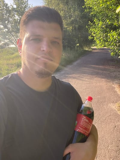 A selfie-style, medium shot captures a man holding a bottle of Coca-Cola in his right arm as he stands on a paved path bordered by grass and trees. The man, with short brown hair and a light beard, wears a dark t-shirt and a gold chain. The sun shines from the left, casting a lens flare across his face and highlighting the green foliage. The path appears to be in a park or a rural area, with the soft light suggesting it is late afternoon. The background shows a treelined path extending into the distance. The scene is outdoors, with the environment suggesting a warm, pleasant day in Yablunivka, Ukraine.