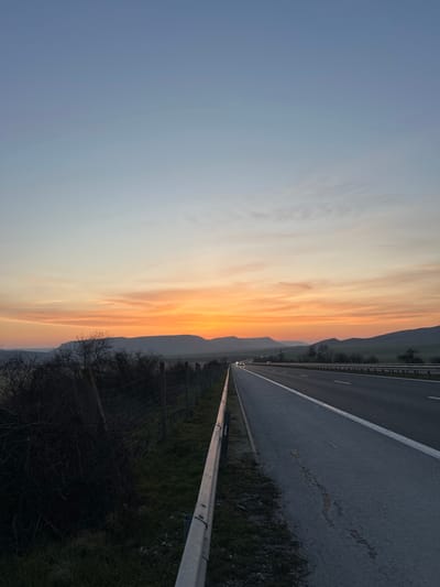 The image depicts a rural or semi-rural roadside scene at dusk or dawn in Mlada Gvardia, Bulgaria. The sky transitions from a light blue at the zenith to vibrant orange and yellow hues at the horizon, indicative of a sunset, with horizontal cloud formations illuminated by the low-angle light. A multi-lane highway, likely an expressway, extends diagonally from the foreground into the distant center, its asphalt surface marked with white lines. Multiple vehicles with activated headlights are visible in the mid-distance on the highway, traveling away from the viewer. Along the left edge of the highway, a galvanized metal crash barrier runs parallel to the road. Beyond the barrier, a strip of green grass precedes a wire mesh fence supported by wooden posts. Dark, leafless deciduous bushes and small trees are situated behind the fence, partially silhouetted against the sky. A chain of dark, undulating hills or mountains forms the distant horizon, silhouetted by the sky. No individuals are present in the scene.