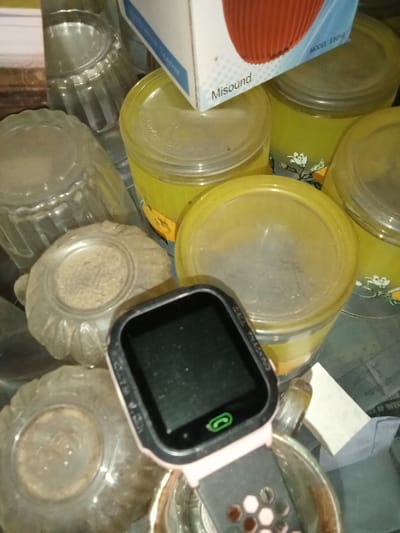The scene, located in Emthanjeni Local Municipality, South Africa, displays a static, cluttered arrangement of domestic objects on a flat surface. A black smartwatch with a dark screen, displaying a green phone icon and with "CALL TRACKING" partially visible on its side, rests on an upright clear glass containing a dark residue. The smartwatch features a pink/light-colored, textured strap. To the left, two inverted clear, ribbed glass bowls are present, each containing a light granular substance. Further left, a clear, ribbed glass lies on its side. Multiple yellow cylindrical plastic containers with clear, dusty lids, some decorated with a white floral pattern, occupy the background and upper right portion of the image. A white and blue cardboard box, labeled "Misound" and partially revealing a red, ribbed object and "MODEL ES010", is positioned on top of some of these yellow containers. A small white rectangular object and a piece of dark fabric are visible behind the glass supporting the smartwatch. Accumulated dust is evident on various surfaces.