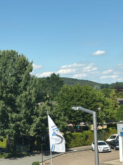The image shows a street scene on a sunny day in Bissendorf, Germany. A white flag with blue and black accents, bearing the text "JIATOHAUS DEPPE", is prominently displayed on the right side of the frame, attached to a flagpole. Behind the flag, a modern street lamp with a grey metallic finish stands tall. In the background, several cars are visible parked on a paved lot, including a white car and a black car. A white van is also parked nearby. The area is surrounded by lush green trees and hedges, with a residential building with a brick facade and a tiled roof in the distance. The sky is a clear blue with a few scattered white clouds. A gentle, tree-covered hill forms the backdrop of the scene. The image appears to be taken from an elevated perspective, looking down on the street and the surrounding environment.